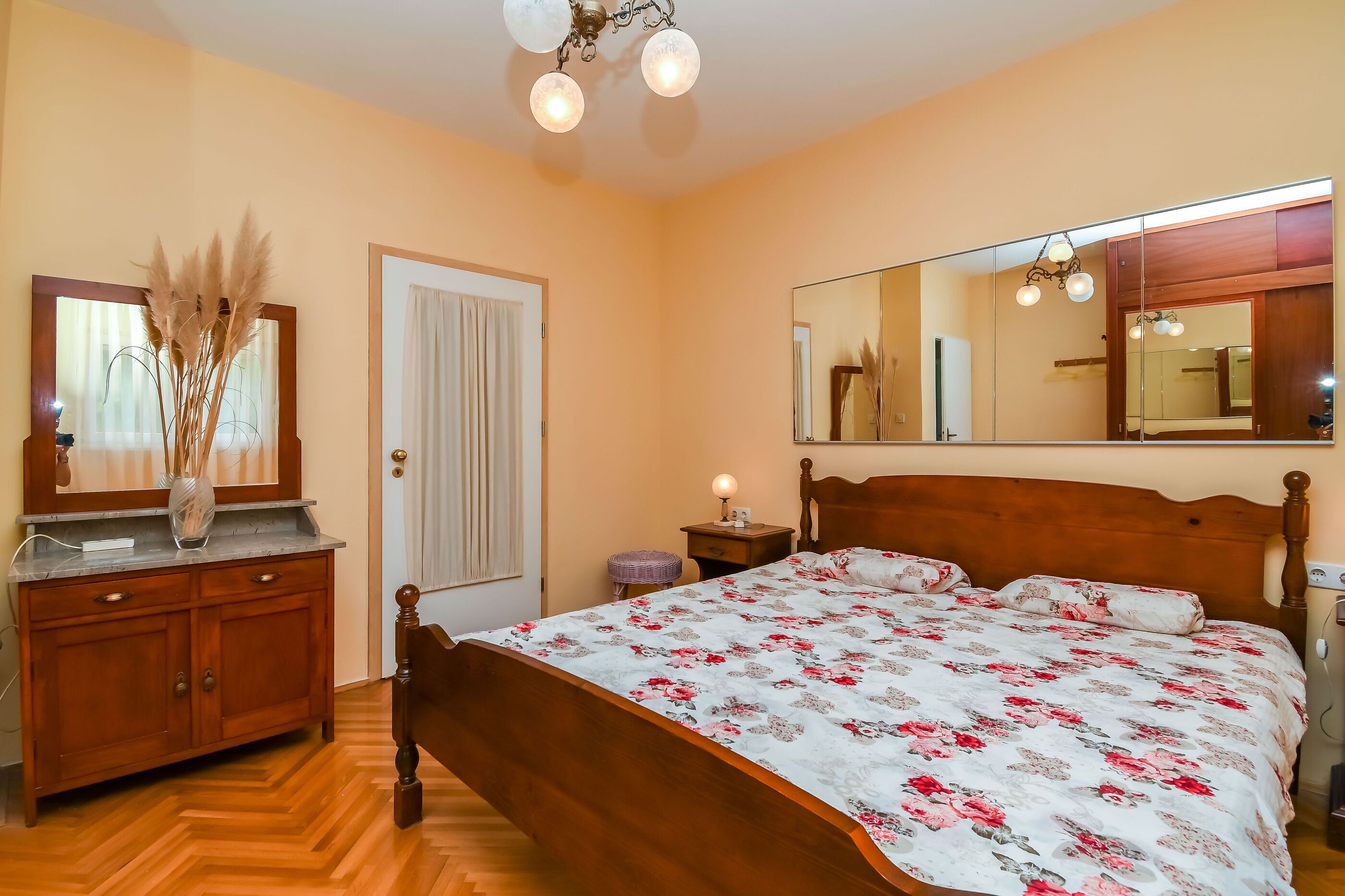 Un dormitorio con una cama de madera, una mesita de noche con lámpara, un aparador, un espejo y un jarrón con plantas secas.