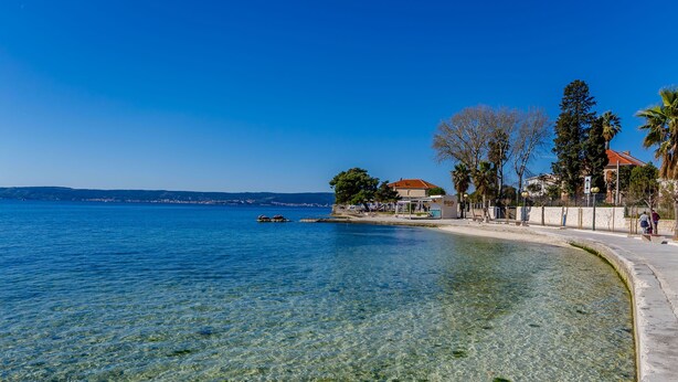 A clear waterfront view with a sandy beach, a small building with a red roof, and a few trees. - Apartments Helios (Kastel Gomilica)