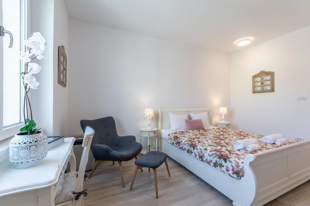 A bedroom with a bed, bedside tables, a chair, a desk, and a window with flowers.