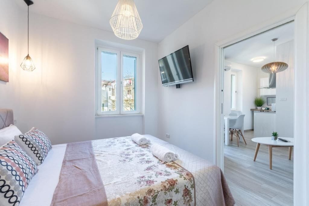 A modern bedroom with a large bed, a TV mounted on the wall, and a view of the outside through a window.