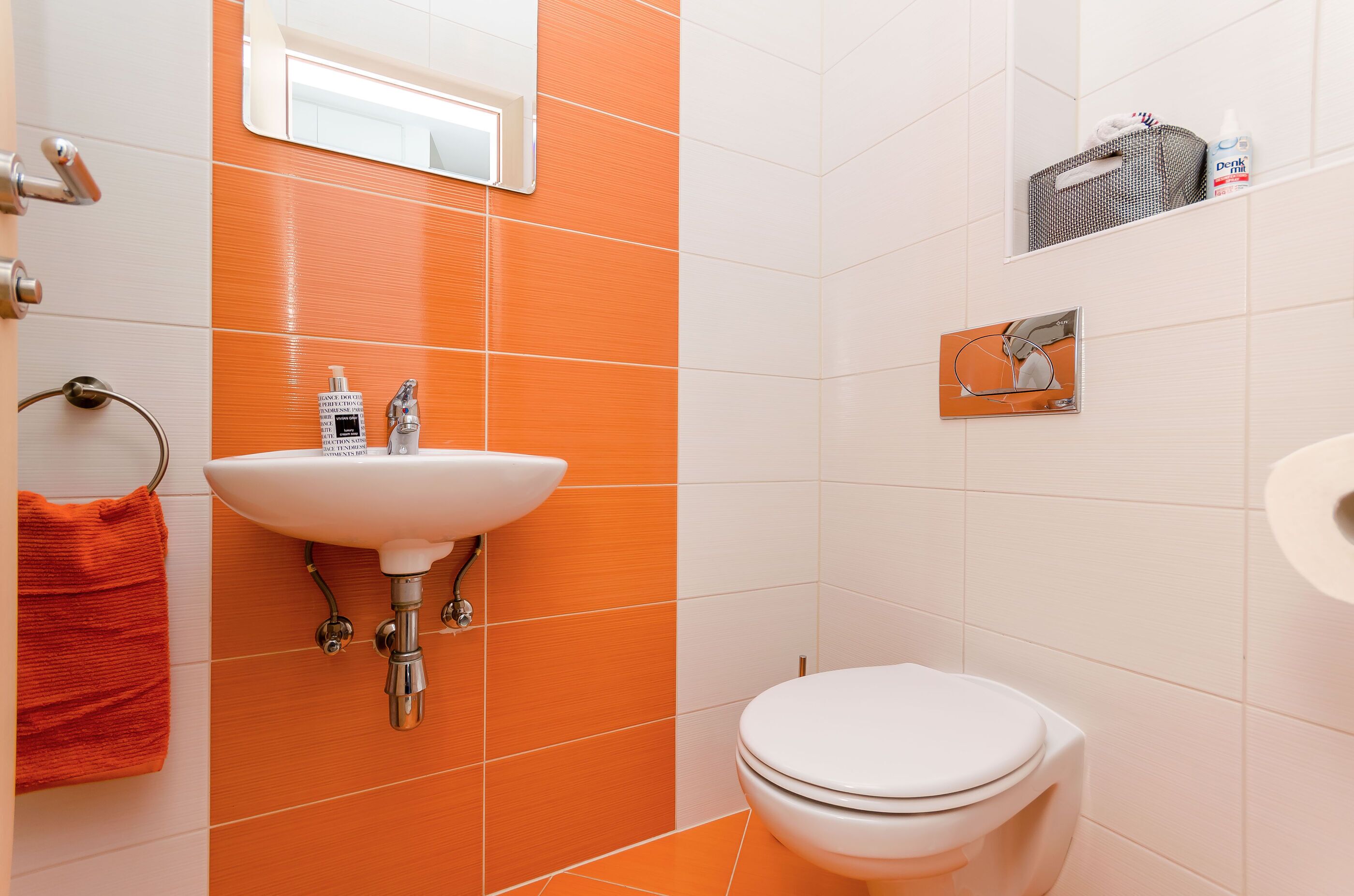 A bathroom with an orange and white tiled wall, a white sink, a toilet, and a mirror.