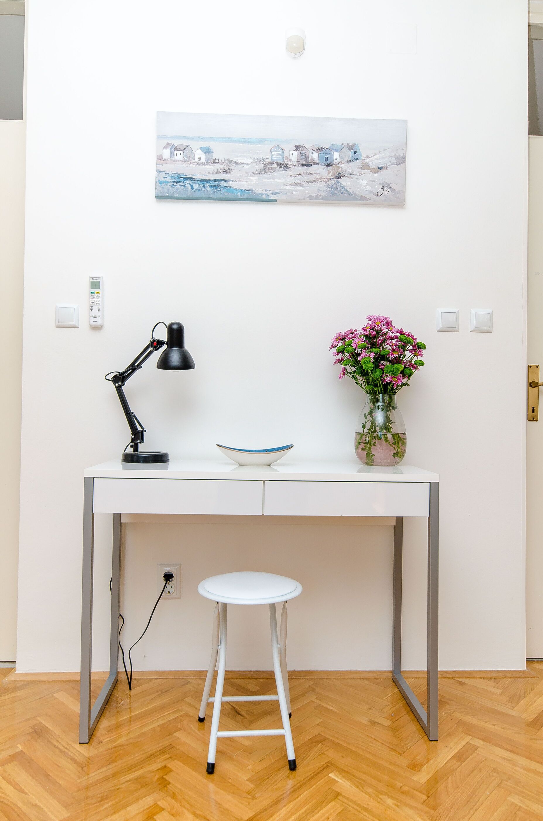 Un bureau blanc avec une lampe de bureau noire, un vase rempli de fleurs roses et un bol bleu.