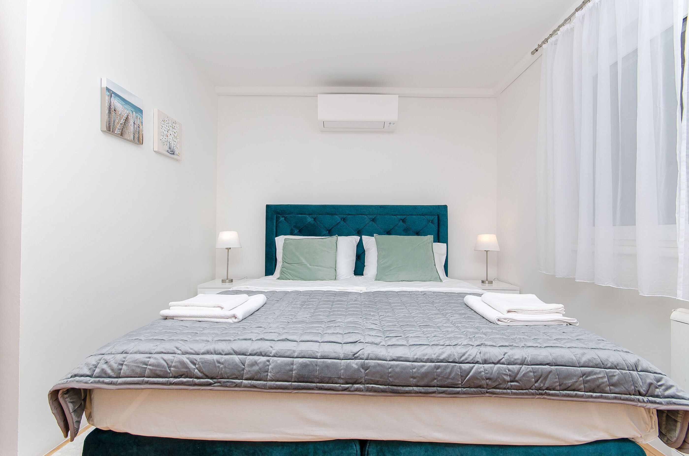 A neatly made bed with a teal headboard, white pillows, and a grey quilt. There are two bedside lamps and a window with sheer curtains.