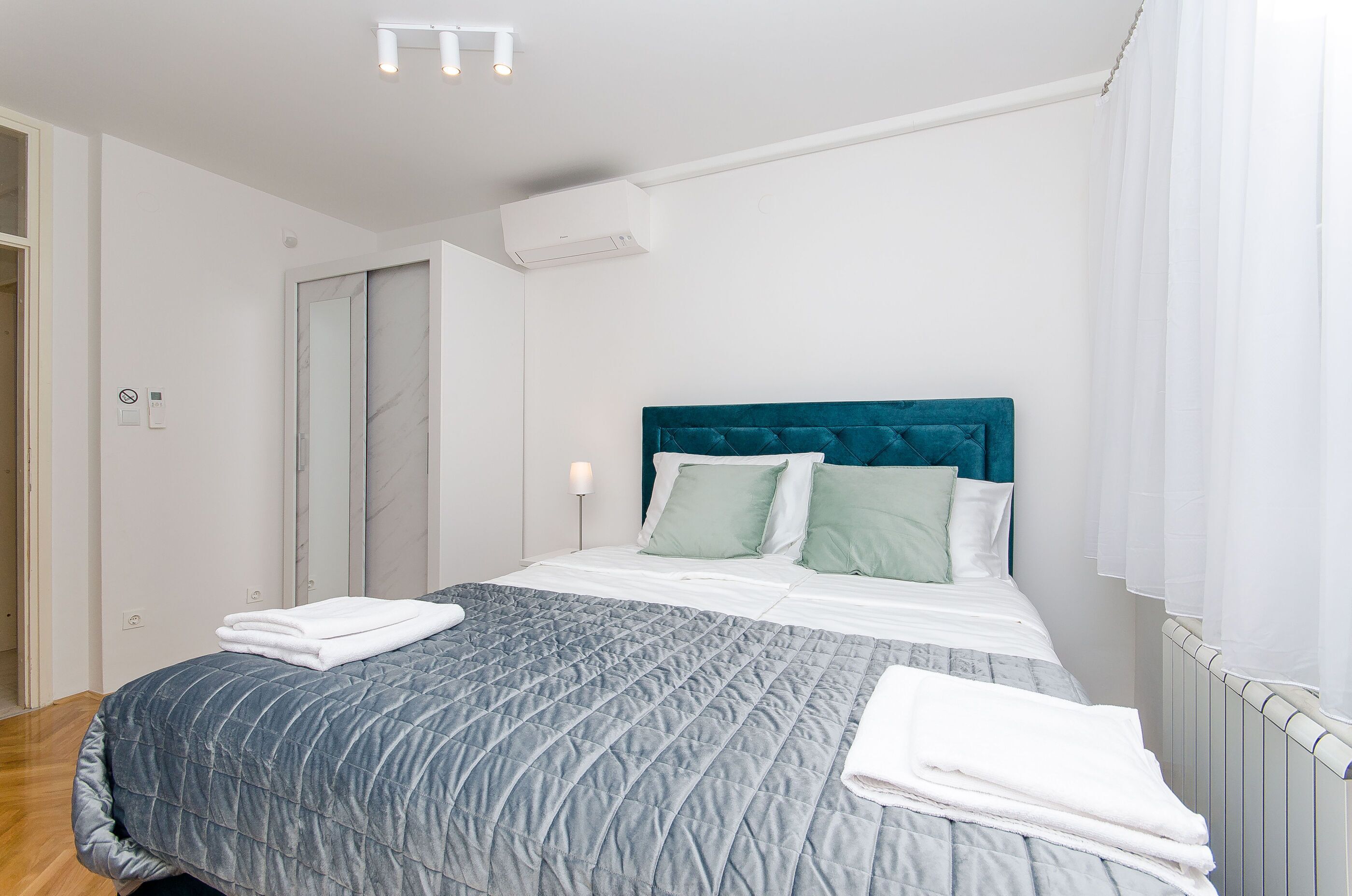 A neatly made bed with a blue quilt, white linens, and two green pillows. There is a white air conditioning unit on the wall, a white radiator, and a white door with a mirror.