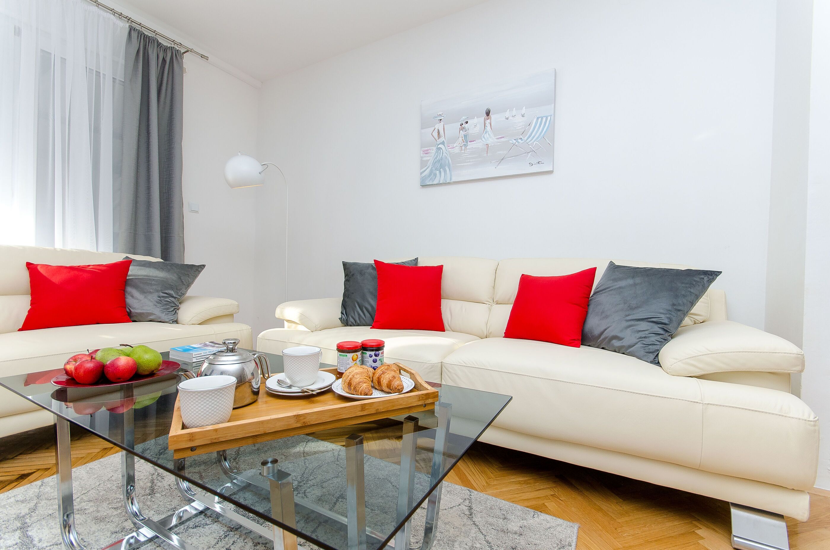 A modern living room with two white sofas, red and grey cushions, a glass coffee table with breakfast items, and a painting on the wall.