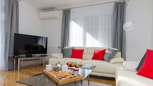 A modern living room with a flat-screen TV, a white sofa with red and gray cushions, a glass coffee table with a tray of food and drinks, and a large window with curtains. - Apartment Gilda Lux (Split)