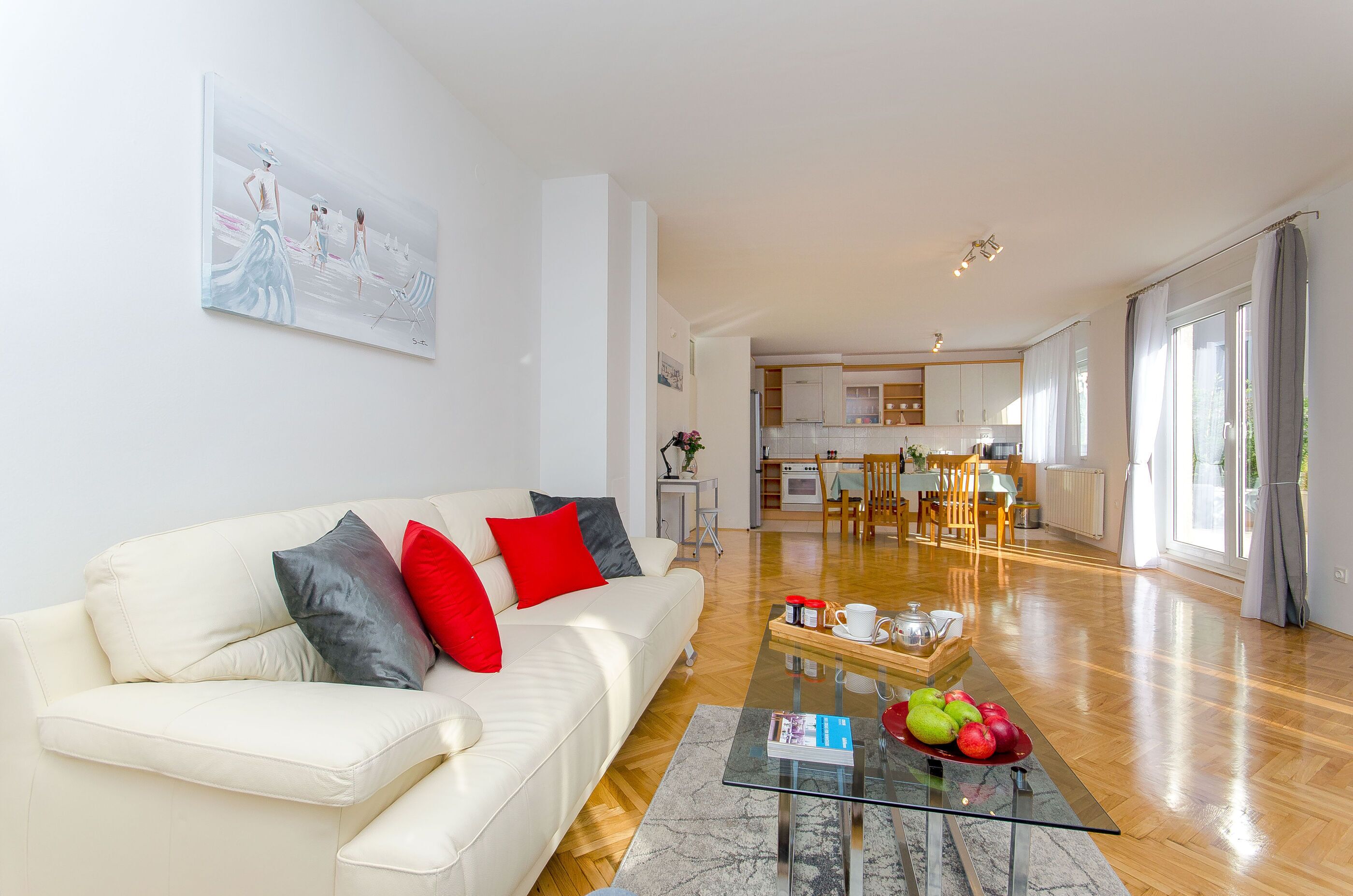 A modern living room with a white sofa, red and grey cushions, a glass coffee table with fruits, and a painting on the wall.