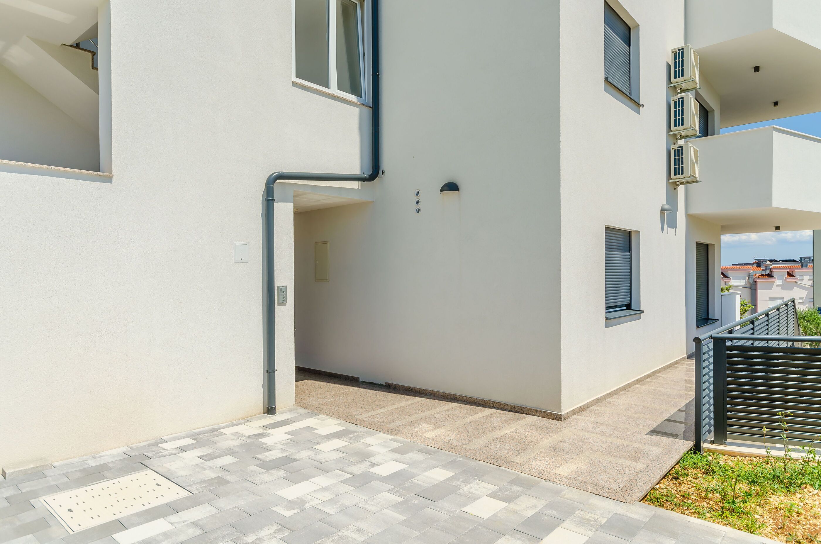 Un bâtiment moderne à la façade blanche et épurée, doté de nombreux balcons et d’une allée piétonne dallée.