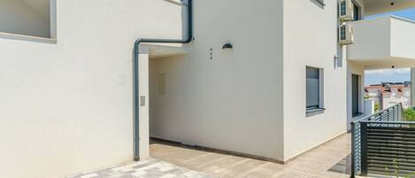 Un bâtiment moderne à la façade blanche et épurée, doté de nombreux balcons et d’une allée piétonne dallée.