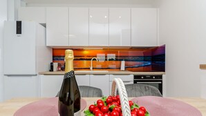 A modern kitchen with white cabinets, a wine bottle, and a basket of cherries on the table. - Apartment Anni (Okrug Gornji)