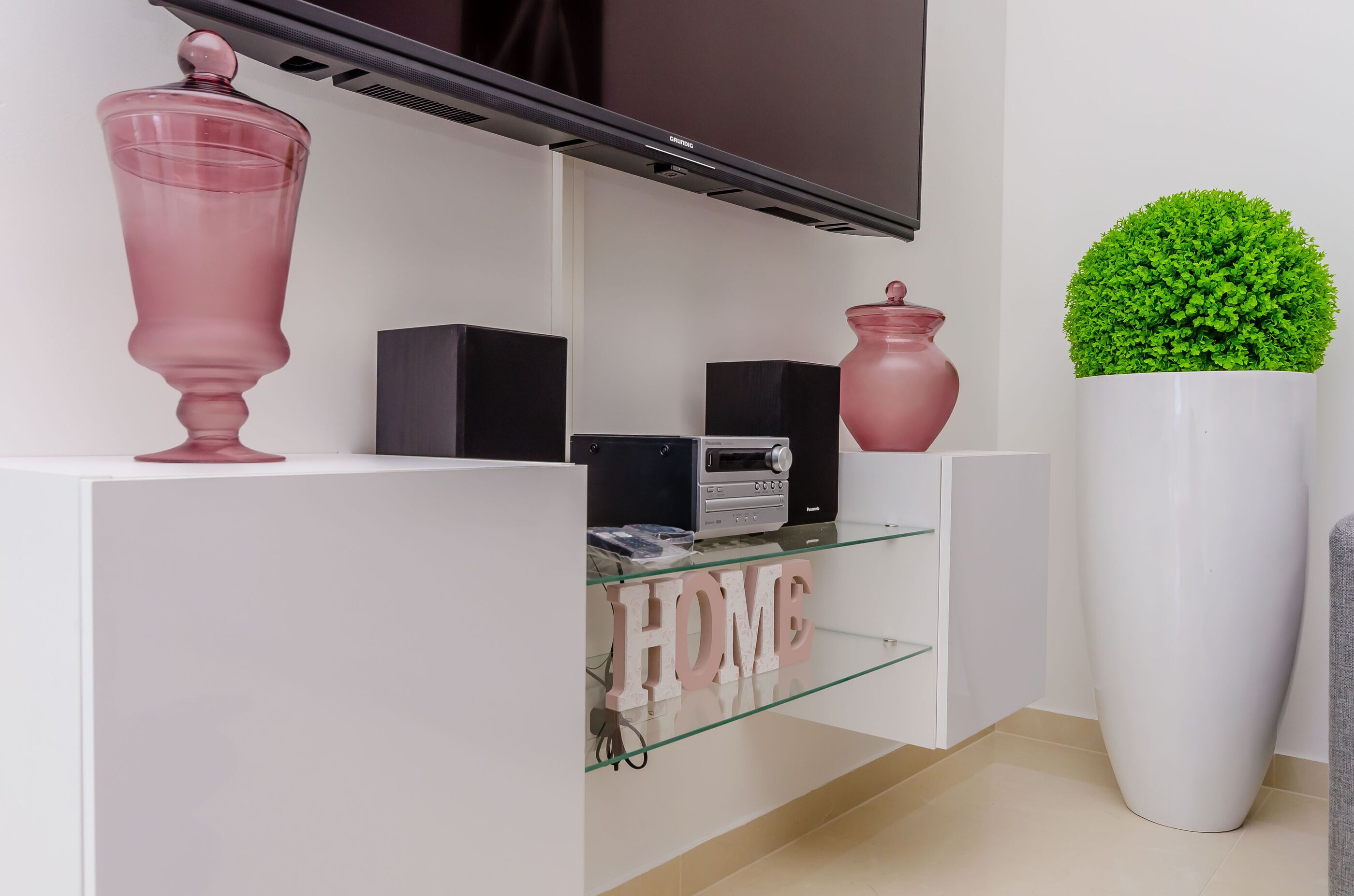 A modern living room with a flat-screen TV mounted on the wall, a glass shelf with decorative items, and a large potted plant.