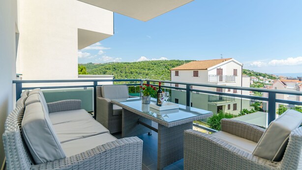 A balcony with wicker furniture, a glass table, and a view of a green landscape and buildings. - Apartment Anni (Okrug Gornji)