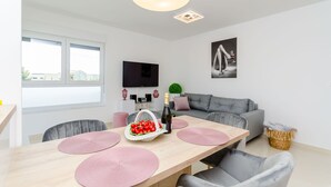 A modern living room with a grey sofa, a wooden dining table with pink placemats, a TV mounted on the wall, and a large window. - Apartment Anni (Okrug Gornji)