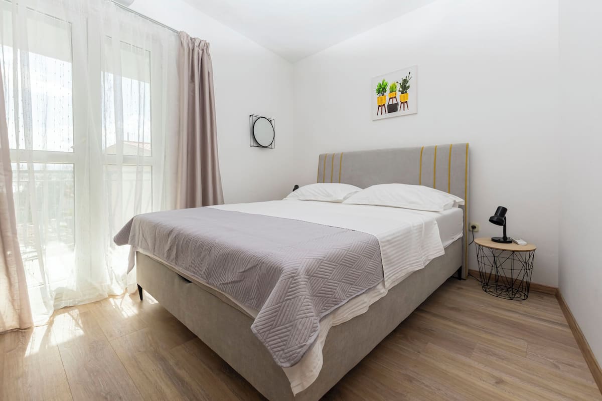 A neatly made bed with a grey blanket, a bedside table with a lamp, and a wall-mounted mirror.