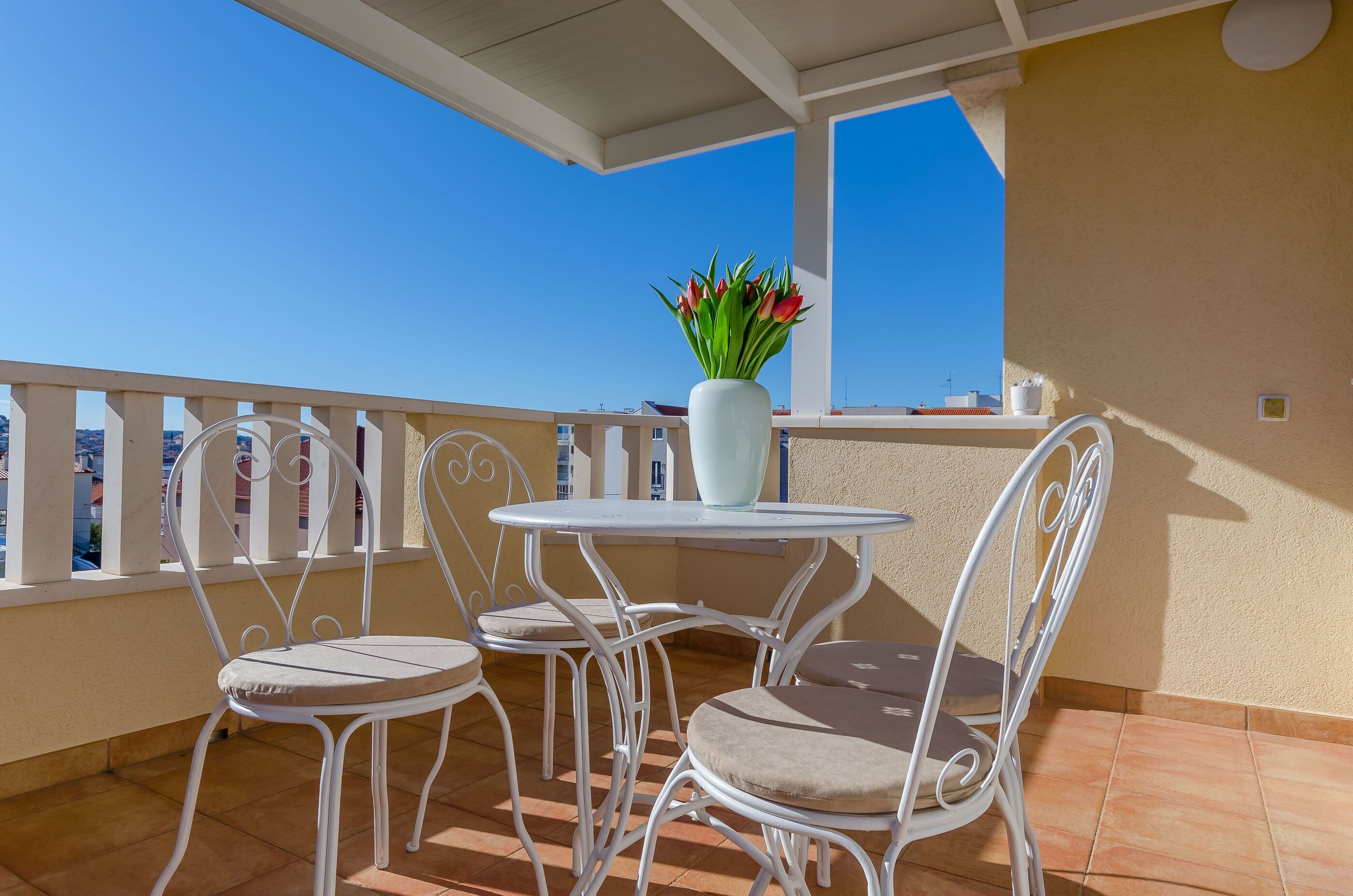 Ein Balkon mit weißen Metallstühlen und einem Tisch, eine weiße Vase mit Blumen und einem strahlend blauen Himmel.