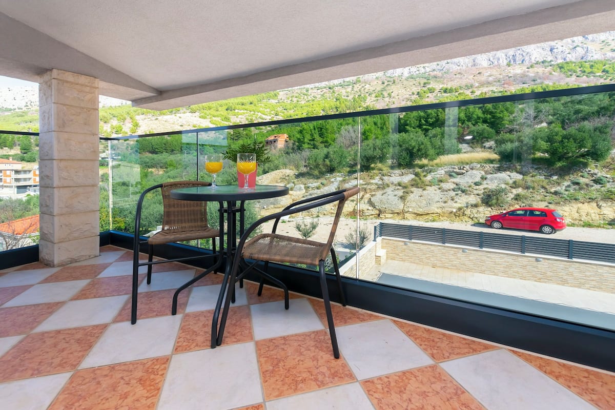 A balcony with a glass railing, two wicker chairs, a small round table with two glasses of orange juice, and a view of a red car on a road with greenery and hills in the background.