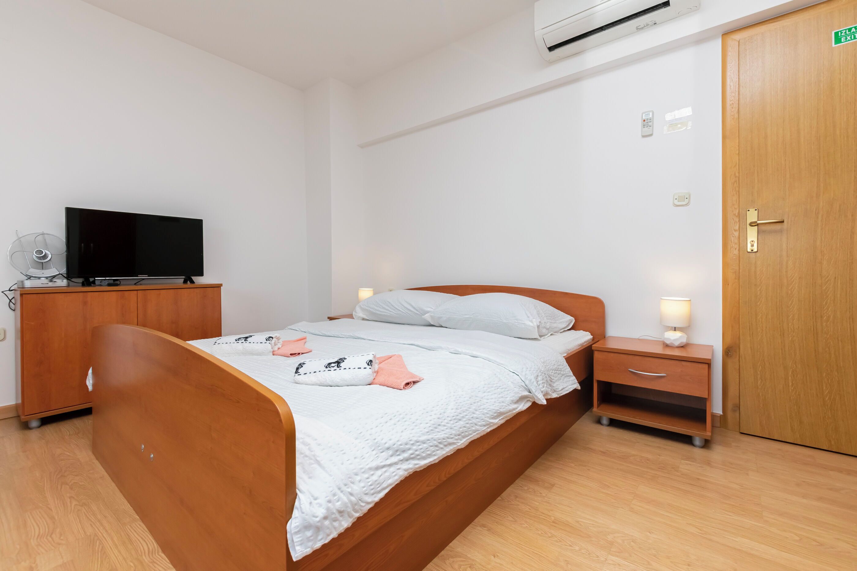 A bedroom with a wooden bed, a bedside table with a lamp, a TV on a wooden cabinet, and an air conditioner on the wall.