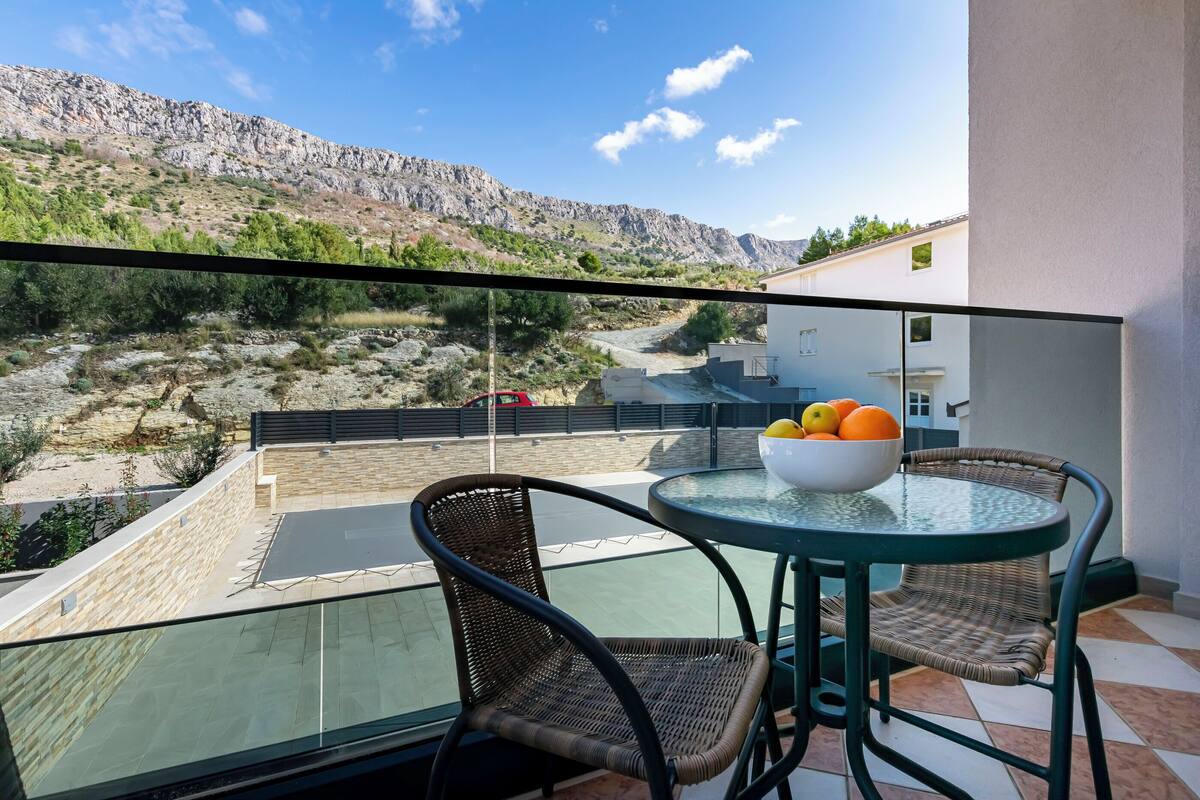 A balcony with wicker chairs and a glass table, overlooking a rocky landscape and a building.