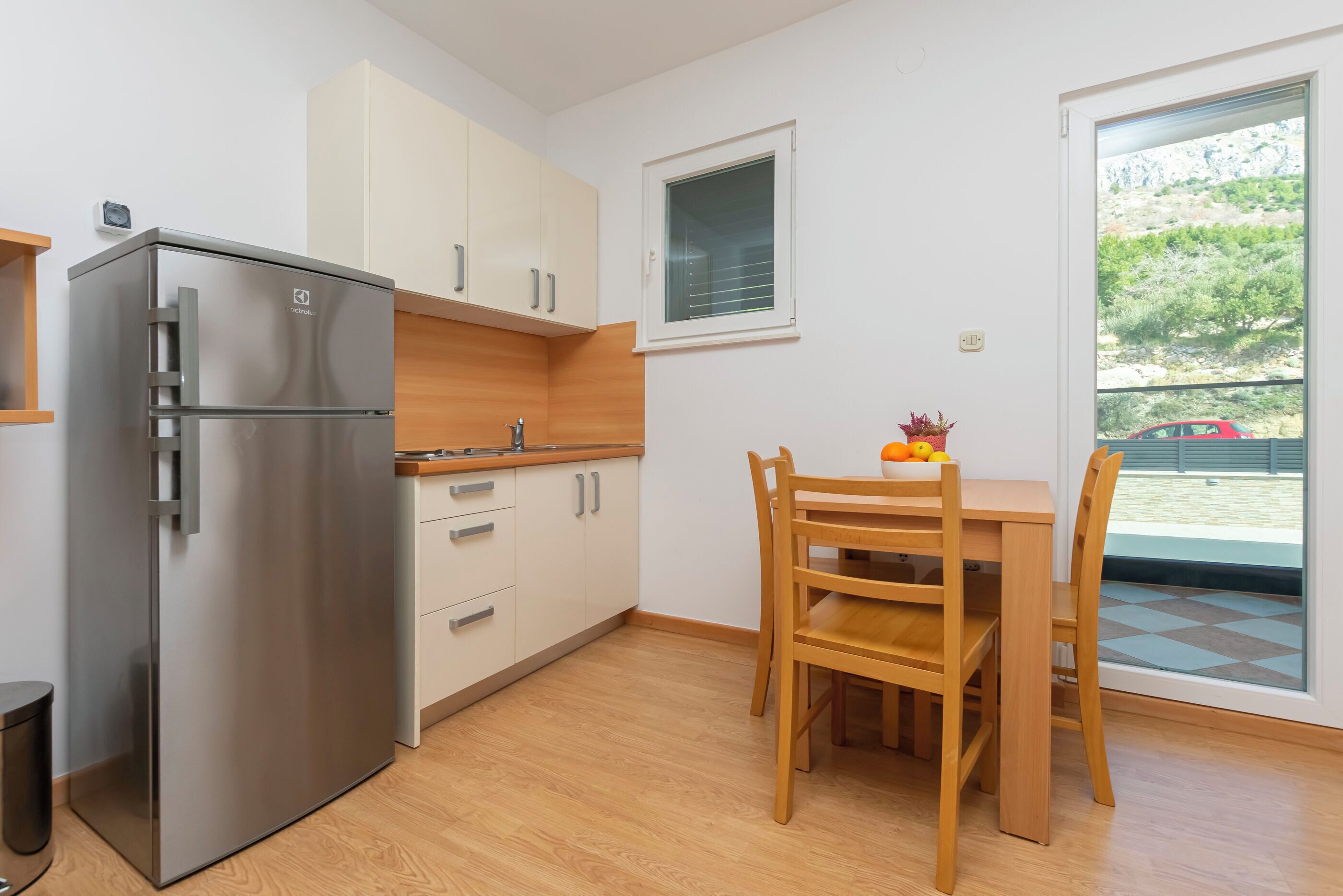 A compact kitchen with a stainless steel refrigerator, wooden cabinets, a dining table with chairs, and a window with a view of a red car and greenery outside.