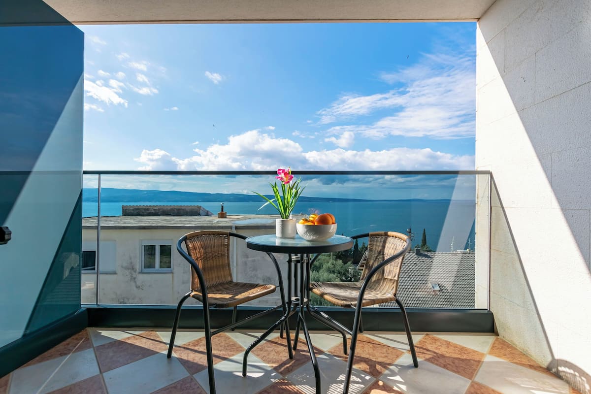 A balcony with a view of the sea, two wicker chairs, a small table with a flower and fruit bowl.