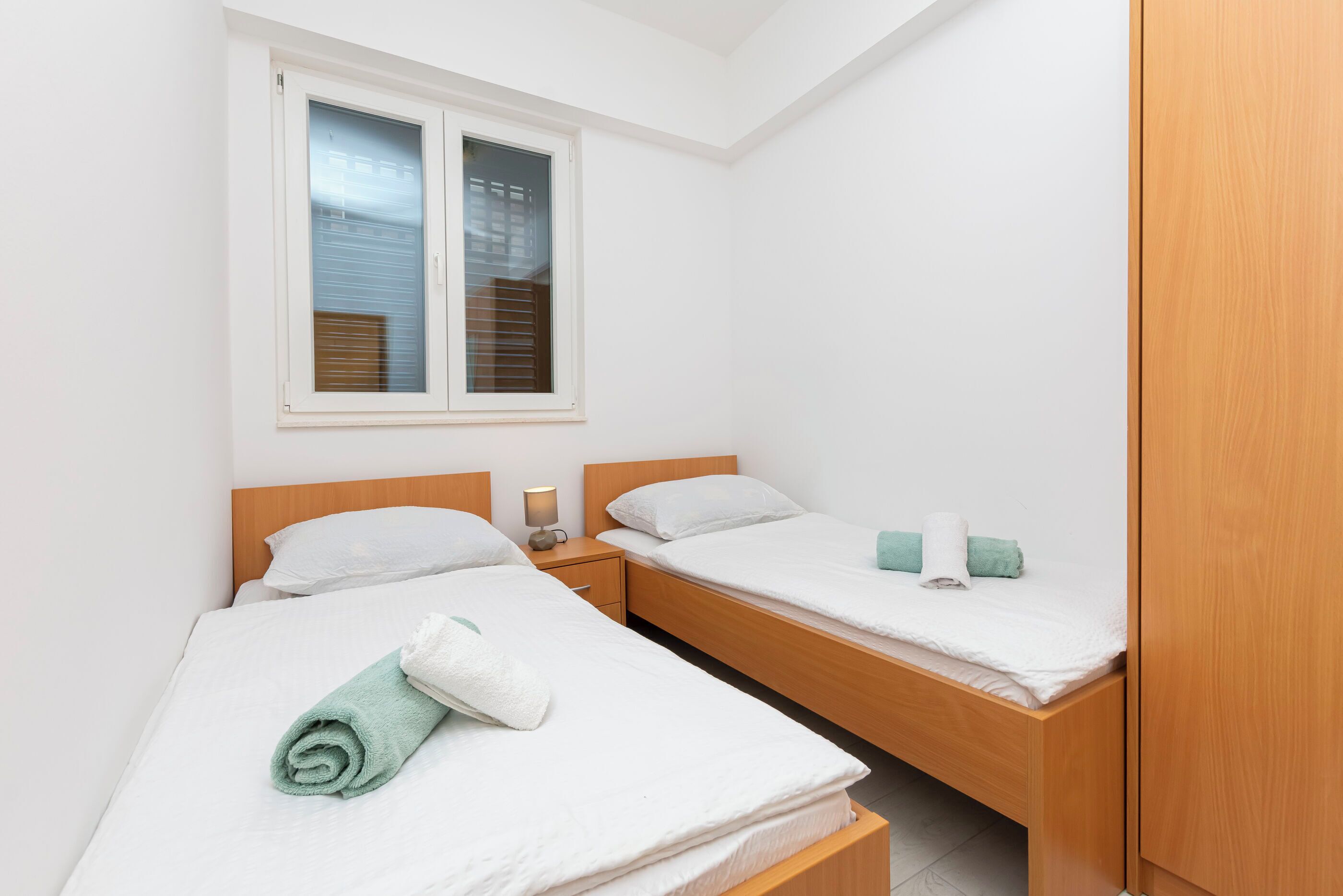A small, clean room with two single beds, a wooden wardrobe, and a window with blinds.