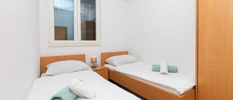A small, clean room with two single beds, a wooden wardrobe, and a window with blinds.