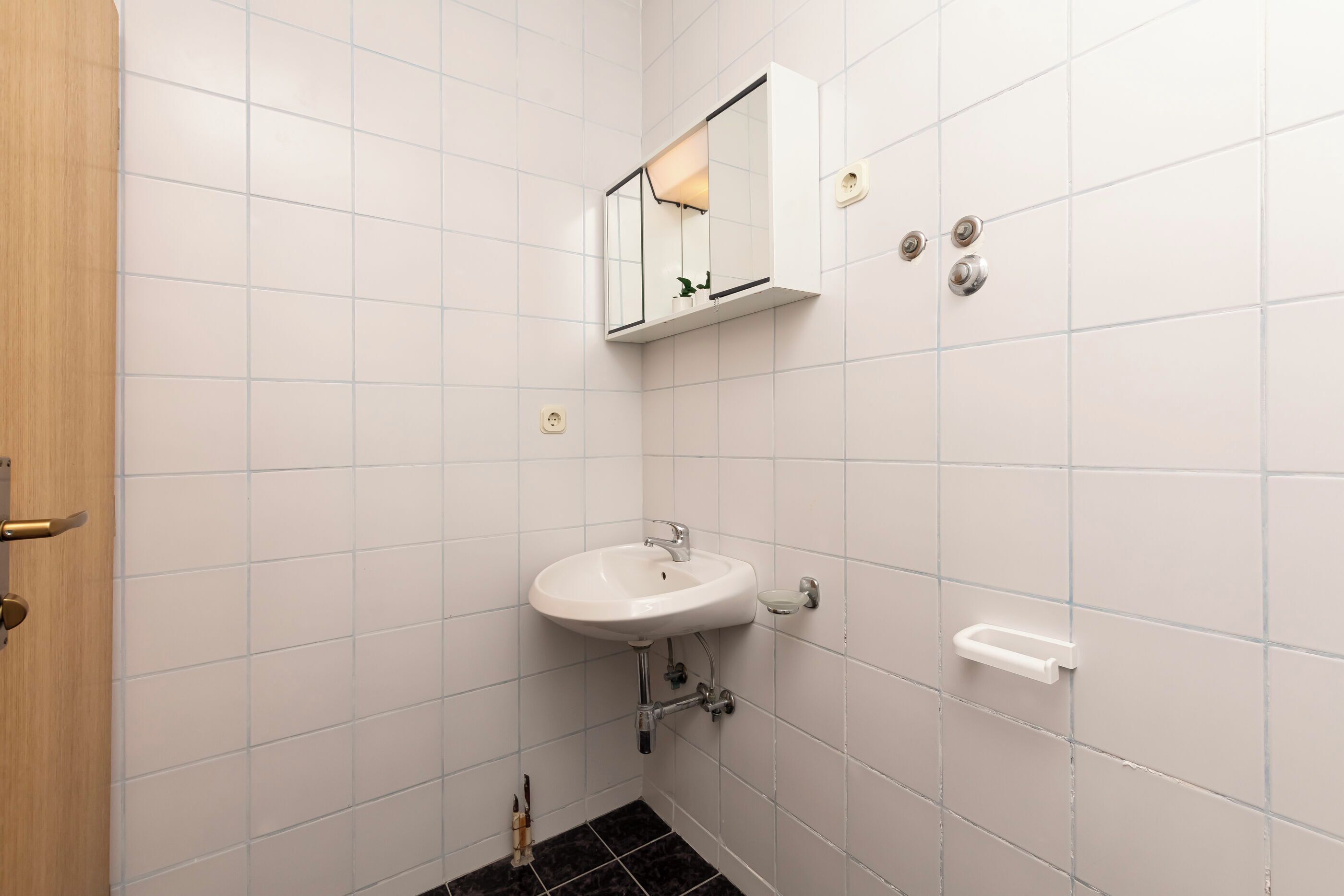 Une salle de bain avec des murs recouverts de carreaux blancs, un lavabo blanc, un miroir et une porte.