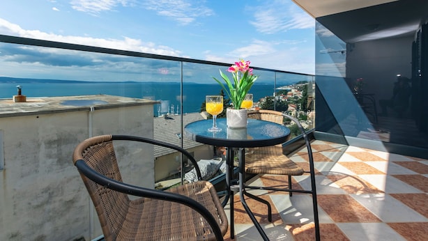 A balcony with wicker chairs and a table, overlooking a scenic view of the sea and distant buildings. - Apartments Villa Milas With Pool (Jesenice)