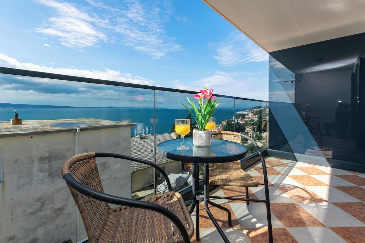 A balcony with wicker chairs and a table, overlooking a scenic view of the sea and distant buildings.