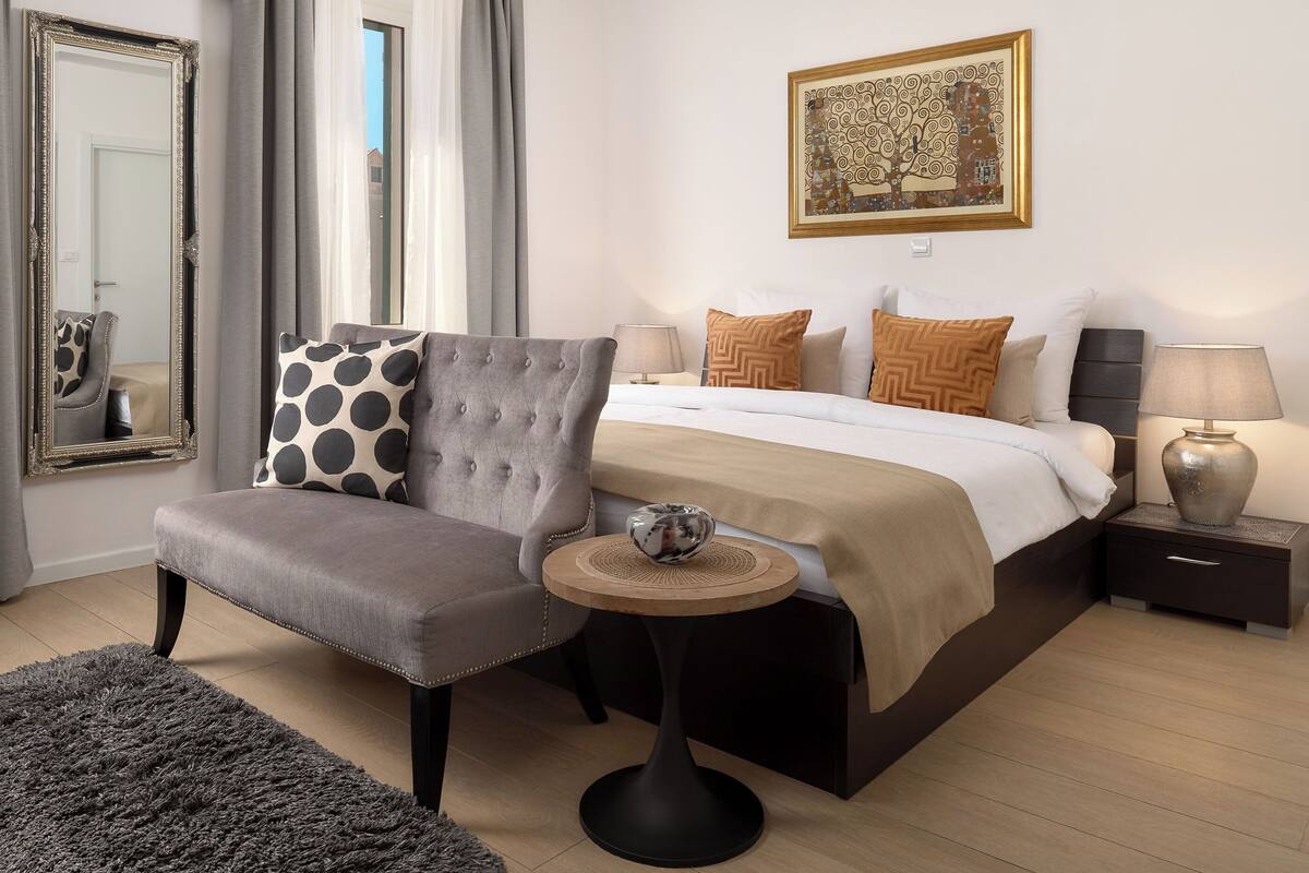 Quarto de hotel com cama, uma cadeira, uma mesa pequena, mesa de cabeceira, abajur, espelho e um quadro na parede.