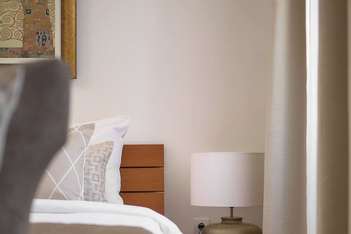Uma cama bem arrumada com edredom branco e almofadas, uma mesa de cabeceira de madeira com abajur, e uma moldura com uma fotografia na parede.