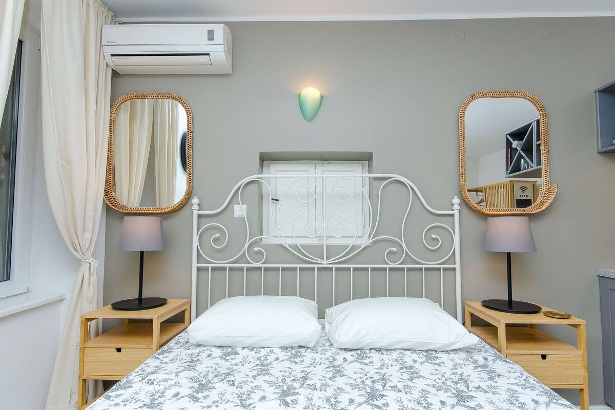 Un dormitorio con una cama, dos mesitas de noche, dos lámparas, dos espejos y un aire acondicionado.