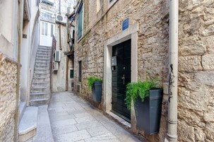 Un bâtiment étroit en pierre, avec un escalier menant à une porte, un portail noir et des plantes en pot au rez-de-chaussée.