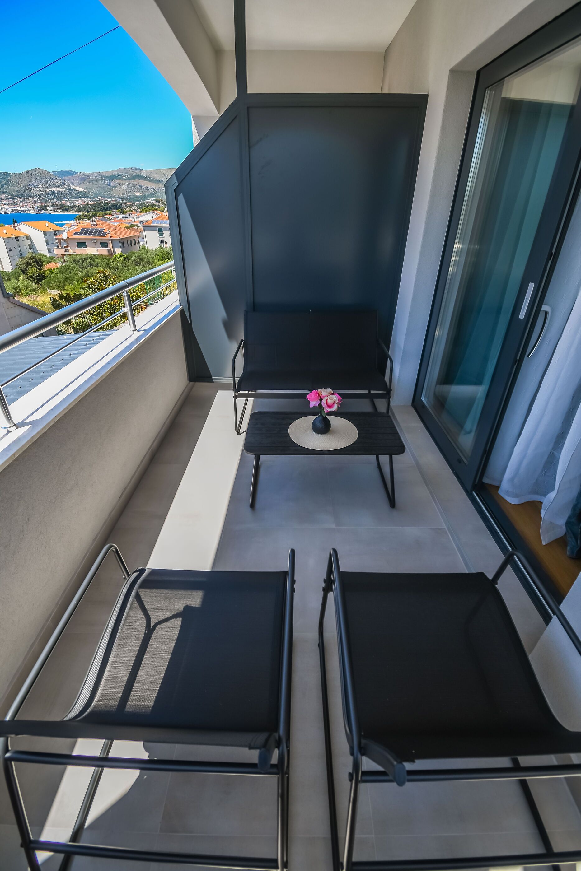 A balcony with two black lounge chairs, a small table with a vase of flowers, and a view of buildings and hills in the background.