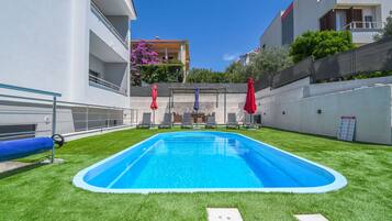 A swimming pool with a surrounding grassy area, a metal railing, and a building with balconies and outdoor furniture.