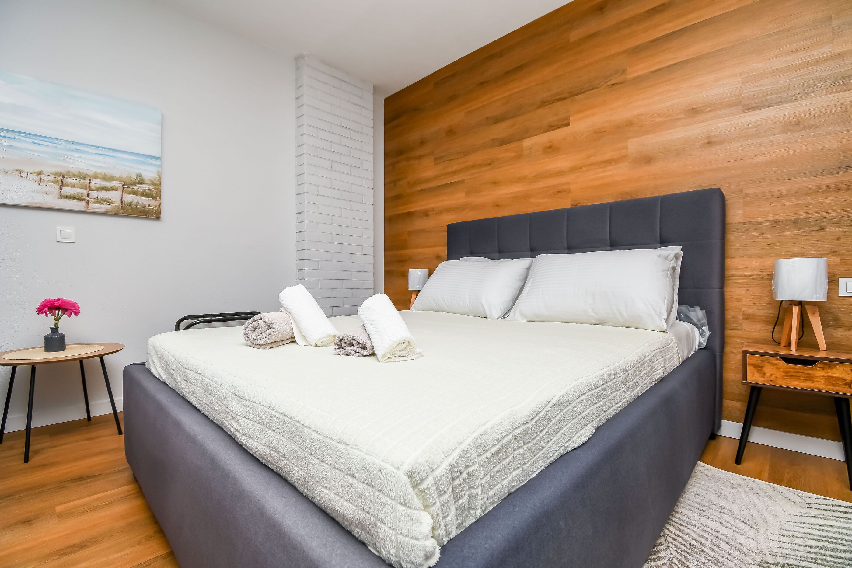 A modern bedroom with a wooden wall, a large bed with white linens, a small round table with a vase of flowers, and a painting of a beach scene.