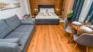 A modern hotel room with a wooden floor, a gray sofa, a neatly made bed with pillows, a dining table with chairs, and a painting of a beach on the wall. - Apartments Neli (Okrug Gornji)