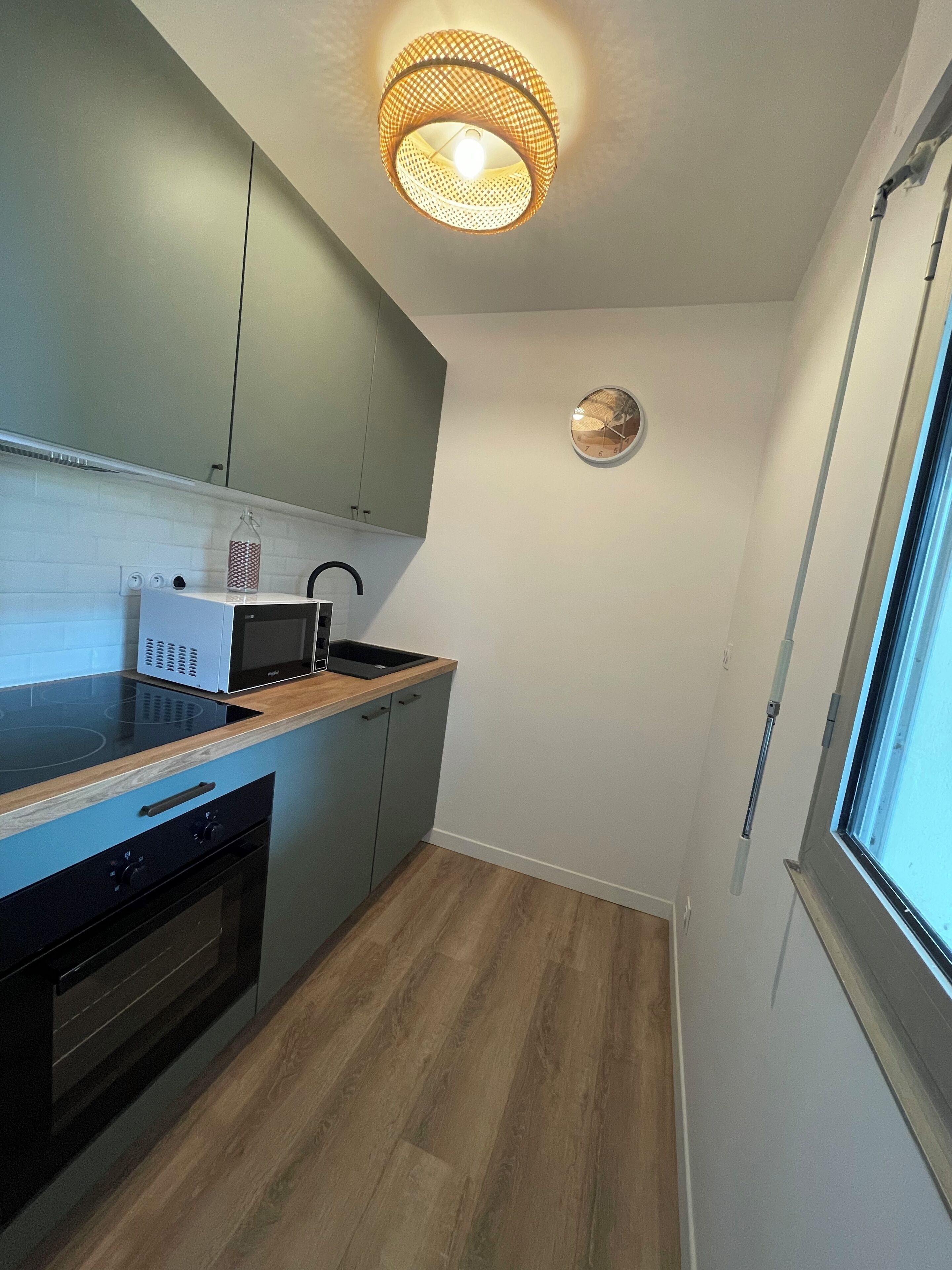 Una cocina compacta con microondas integrado, un lavamanos y una estufa. Cuenta con una encimera de madera, una lámpara colgante y un reloj de pared.