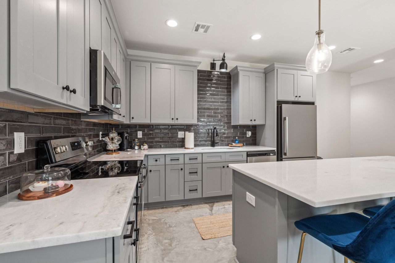 A modern kitchen with grey cabinets, a black tiled backsplash, stainless steel appliances, and a marble countertop.