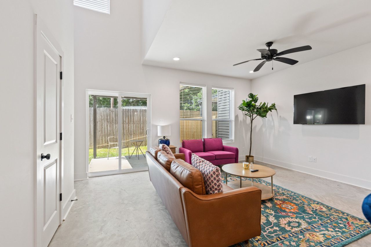 A spacious living room with a leather sofa, a pink armchair, and a coffee table. There is a large window with blinds, a ceiling fan, and a flat-screen TV mounted on the wall.