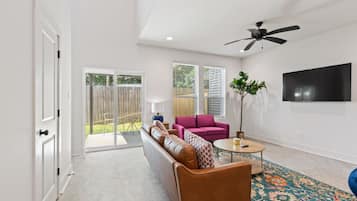 A spacious living room with a leather sofa, a pink armchair, and a coffee table. There is a large window with blinds, a ceiling fan, and a flat-screen TV mounted on the wall.
