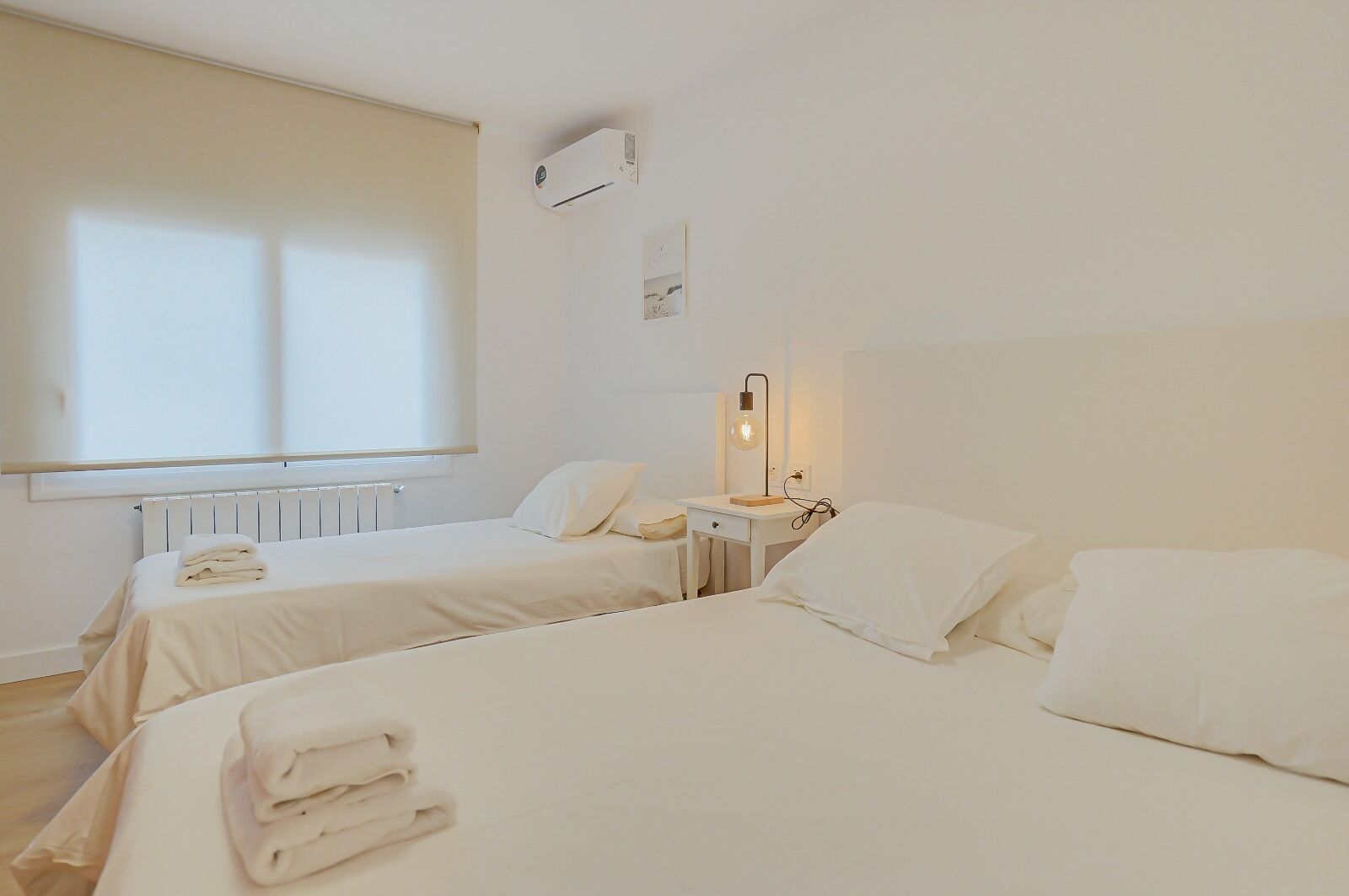 Hotellihuone, jossa on kaksi sänkyä, yöpöytä lampulla, patteri ja ikkuna, jossa on sälekaihtimet.