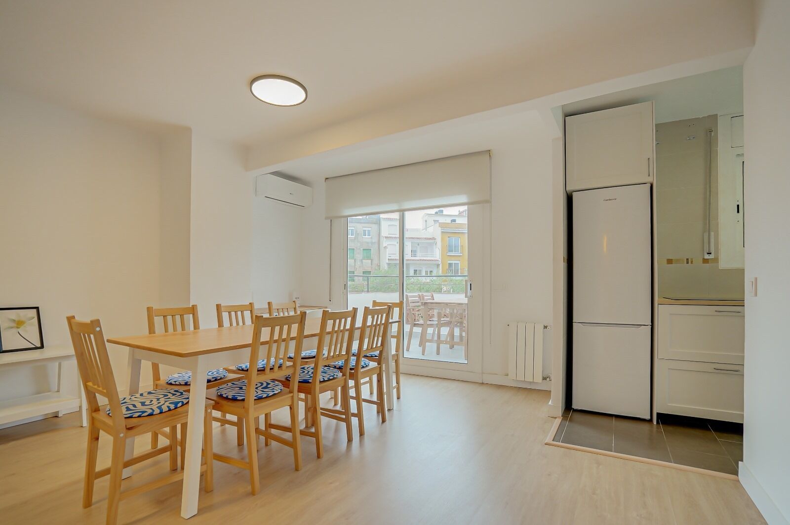 Un comedor con una mesa y sillas de madera, una cocina con refrigerador y vistas a edificios a través de un ventanal grande.