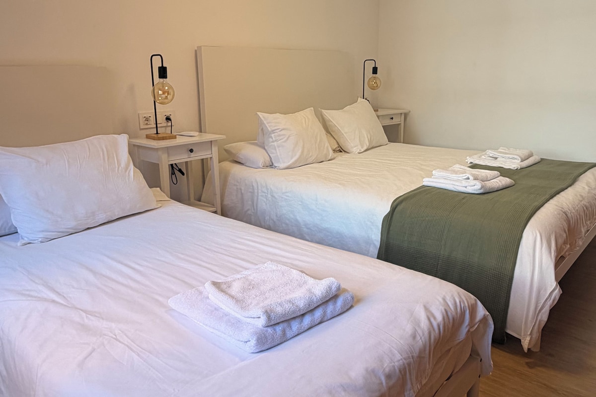 Habitación de hotel con dos camas, cada una con sábanas y almohadas blancas, una mesita de noche pequeña con lámpara y una manta verde sobre una de las camas.