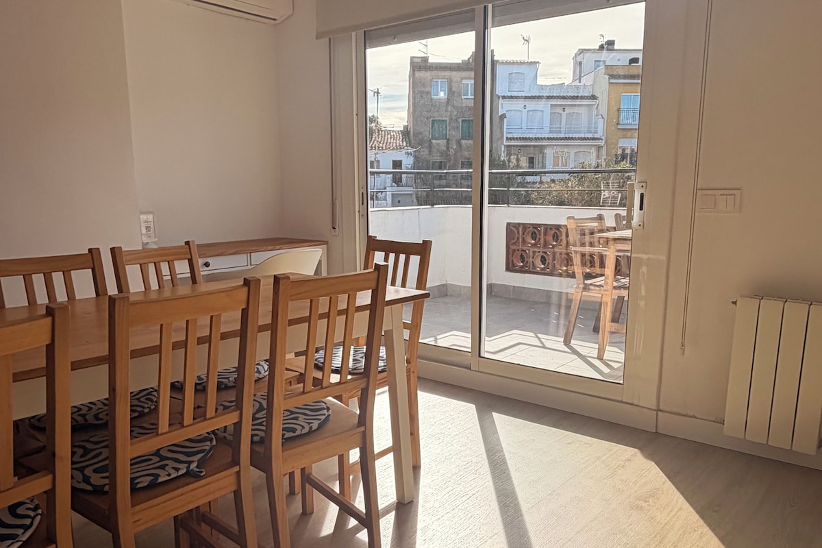 Un coin repas avec une table et des chaises en bois, une grande porte-fenêtre coulissante donnant sur un balcon, et un climatiseur blanc fixé au mur.