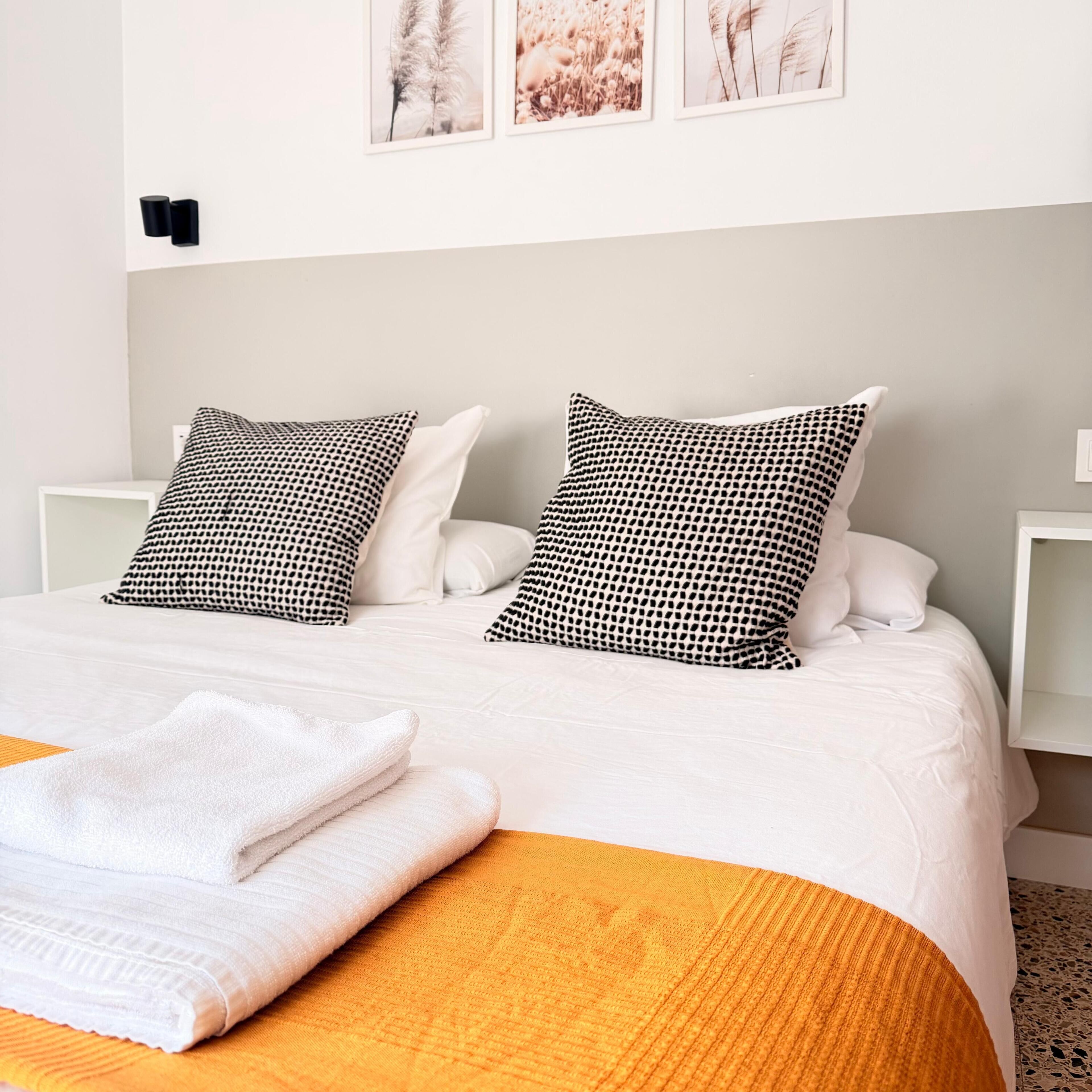 Een net opgemaakt bed met witte beddengoed en twee zwart-wit gekleurde kussens, een opgevouwen witte handdoek en een gele decoratieve plaid.
