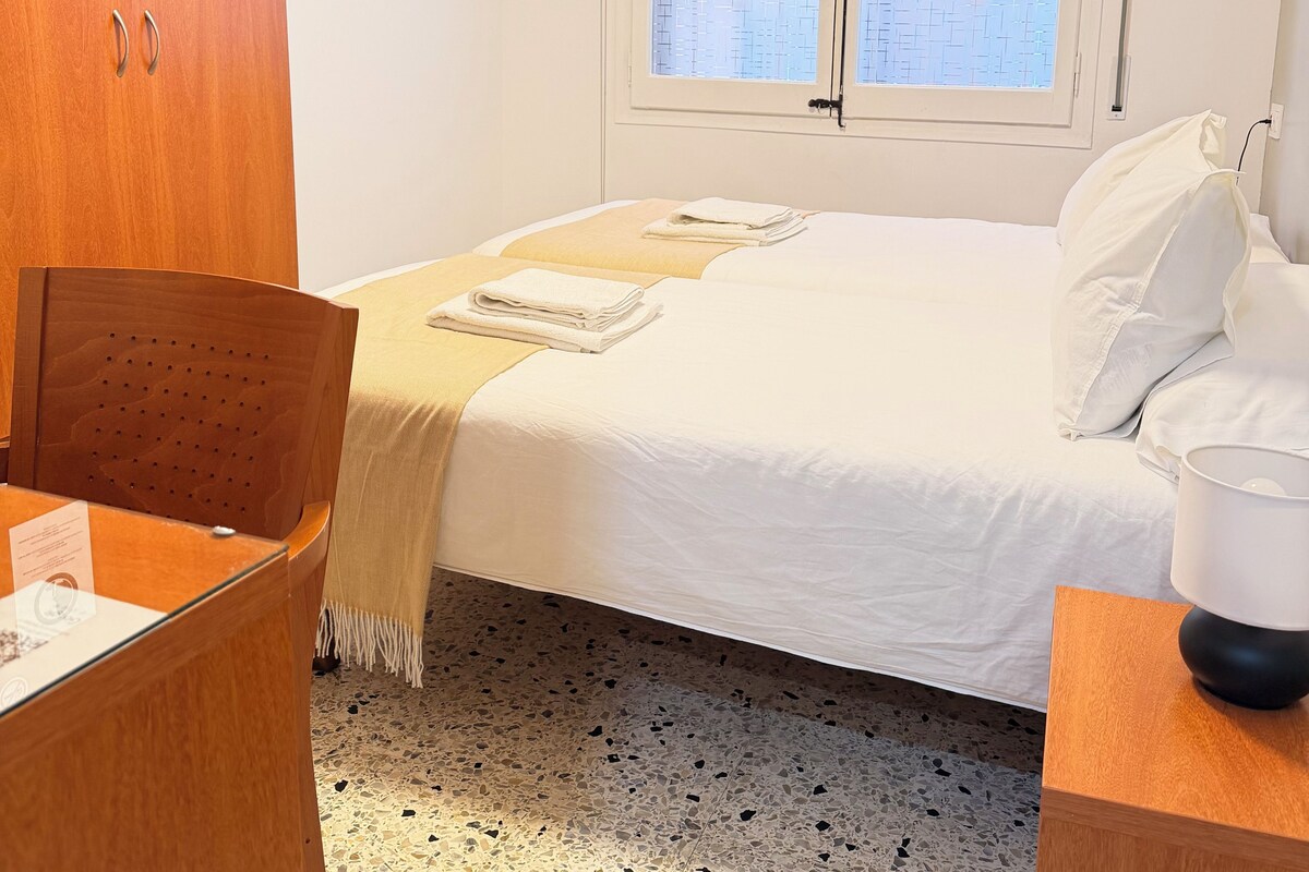 Een hotelkamer met een bed, een bureau, een stoel, een aan de muur bevestigde televisie en een raam met jaloezieën.