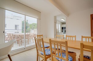 Un comedor con una mesa grande y sillas, un muro de vidrio corredizo que da a un balcón exterior, y una cocina visible a través de una puerta abierta.