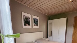 Makuuhuoneessa on puinen katto, valkoisilla tekstiileillä varustettu sänky, yöpöytä, vaatekaappi ja kaksi kehystettyä kuvaa seinällä.
