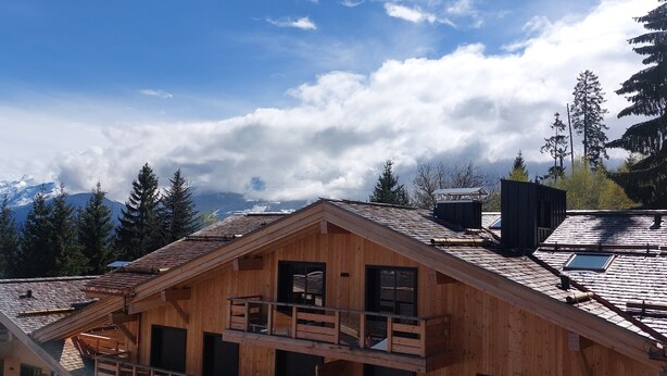 A wooden cabin with a balcony, a small attached structure, and a view of mountains in the background.